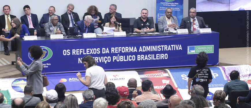 A imagem mostra a audiência pública realizada na Câmara dos Deputados, em Brasília. Na mesa principal, representantes de entidades sindicais e parlamentares discutem. Sobre a imagem, o texto diz: “Fenajud reforça resistência nacional à Reforma Administrativa na Câmara dos Deputados”.