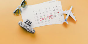 Arte em tons de laranja com uma mesa sobre a qual estão um calendário, uma câmera fotográfica, um pequeno avião de brinquedo e óculos de sol. Algumas datas estão circuladas em vermelho.