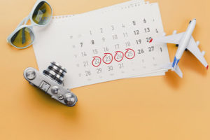 Arte em tons de laranja com uma mesa sobre a qual estão um calendário, uma câmera fotográfica, um pequeno avião de brinquedo e óculos de sol. Algumas datas estão circuladas em vermelho.