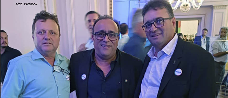 Foto em que há três homens de pé, abraçados e com roupas sociais olhando para a câmera. Ao centro está o presidente da Feserv-Minas, Hely Aires.
