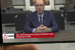 Montagem digital de uma mão segurando um smartphone, na tela do aparelho está uma cena do pronunciamento do Des. José Arthur Filho, um homem branco, calvo de olhos azuis, vestido em traje social.