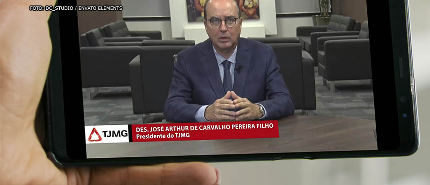 Montagem digital de uma mão segurando um smartphone, na tela do aparelho está uma cena do pronunciamento do Des. José Arthur Filho, um homem branco, calvo de olhos azuis, vestido em traje social.