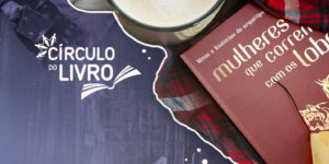 Banner do Círculo do Livro com o livro Mulheres que correm com os lobos, de Clarissa Pinkola Estés, ao lado de uma xícara de café sobre uma manta xadrez. Ao fundo, há uma superfície de madeira escura e o logotipo “Círculo do Livro”.
