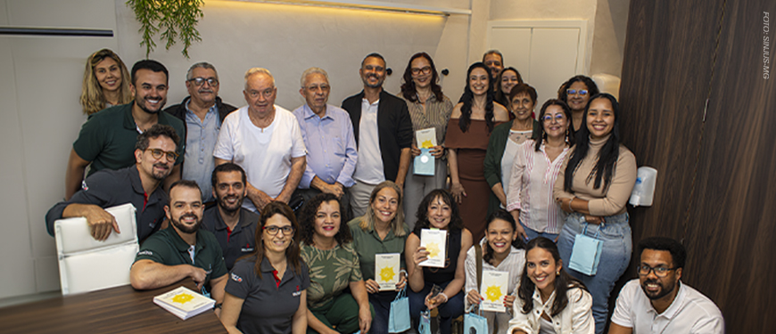 Imagem acessível: A imagem mostra um grupo de pessoas reunido durante a inauguração do Espaço Terapêutico Nicolau Prímola, na sede do SINJUS-MG. Em destaque, o título diz “SINJUS inaugura Espaço Nicolau Prímola para ampliar ações de saúde e acolhimento”. Na base da imagem, aparece a logomarca do SINJUS.
