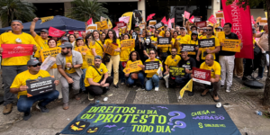 Manifestação de servidores em frente à sede do TJMG. O grupo, majoritariamente vestido de amarelo, segura cartazes cobrando direitos como Data-Base, Promoção Vertical e combate à terceirização. Algumas pessoas usam máscaras de terror em referência ao Halloween. Em primeiro plano, uma grande faixa no chão traz a frase “Direitos em dia ou protesto todo dia”, acompanhada de abóboras, morcegos e elementos de Halloween.