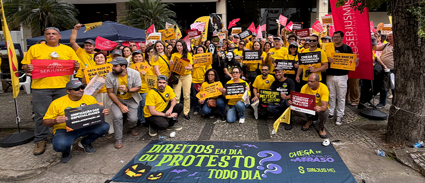 Manifestação de servidores em frente à sede do TJMG. O grupo, majoritariamente vestido de amarelo, segura cartazes cobrando direitos como Data-Base, Promoção Vertical e combate à terceirização. Algumas pessoas usam máscaras de terror em referência ao Halloween. Em primeiro plano, uma grande faixa no chão traz a frase “Direitos em dia ou protesto todo dia”, acompanhada de abóboras, morcegos e elementos de Halloween.