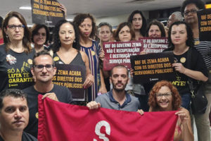 Fotografia com a mobilização dos servidores públicos na ALMG. As pessoas estão sérias, usando roupas escuras e levantam cartazes com frases sobre o RRF, mais abaixo está a bandeira vermelha do SINJUS-MG.