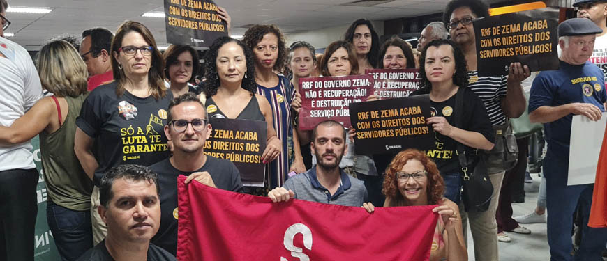 Fotografia com a mobilização dos servidores públicos na ALMG. As pessoas estão sérias, usando roupas escuras e levantam cartazes com frases sobre o RRF, mais abaixo está a bandeira vermelha do SINJUS-MG.