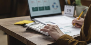 A imagem mostra uma pessoa usando uma calculadora e fazendo anotações sobre uma mesa, com um notebook aberto ao fundo exibindo gráficos e um pequeno calendário destacando o dia 19.