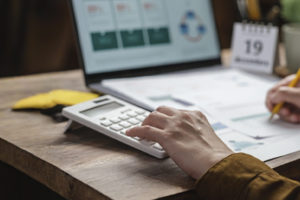 A imagem mostra uma pessoa usando uma calculadora e fazendo anotações sobre uma mesa, com um notebook aberto ao fundo exibindo gráficos e um pequeno calendário destacando o dia 19.