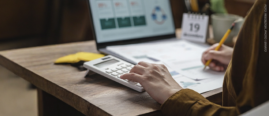 A imagem mostra uma pessoa usando uma calculadora e fazendo anotações sobre uma mesa, com um notebook aberto ao fundo exibindo gráficos e um pequeno calendário destacando o dia 19.