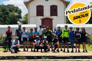 Esta imagem mostra um grupo de ciclistas com suas bicicletas. Eles estão ao ar livre, em frente a uma igreja, com árvores ao redor. No canto superior, há um logotipo com o texto "Pedalando com o SINJUS".