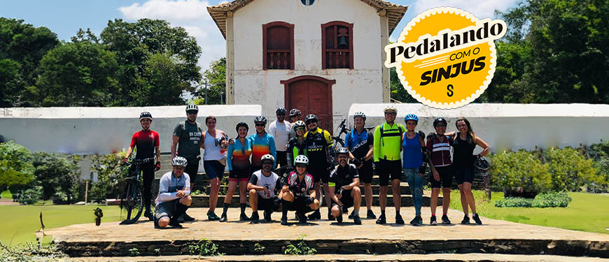 Esta imagem mostra um grupo de ciclistas com suas bicicletas. Eles estão ao ar livre, em frente a uma igreja, com árvores ao redor. No canto superior, há um logotipo com o texto "Pedalando com o SINJUS".