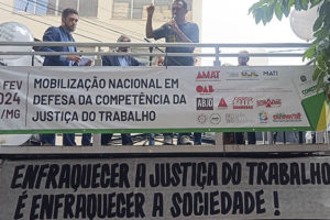 Jonas Araújo está em cima de um carro de som, com o punho erguido. Na placa estendida a grade de proteção, lê-se: Mobilização Nacional em defesa da Justiça do Trabalho.