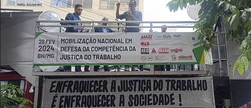 Jonas Araújo está em cima de um carro de som, com o punho erguido. Na placa estendida a grade de proteção, lê-se: Mobilização Nacional em defesa da Justiça do Trabalho.