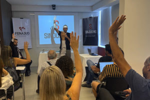 Imagem Acessível: A imagem retrata uma assembleia presencial realizada pelo SINJUS-MG, com filiados reunidos em uma sala votando com as mãos levantadas. Ao fundo, um diretor está em frente a um telão com o logotipo do sindicato, conduzindo a votação. Também há banner da FENAJUD disposto na lateral. O texto em destaque informa: “Filiados aprovam novas ações de luta e contas do Sindicato”. O ambiente é iluminado e participativo, evidenciando o caráter democrático da AGE e AGO.
