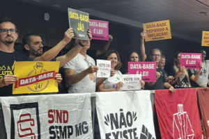 A imagem registra um momento de aprovação da luta sindical na ALMG, com servidores e representantes sindicais segurando cartazes coloridos com frases como “Data-Base Já!”, “Data-Base é Lei!” e “Vote Sim”. Na sacada do plenário, há faixas de diversos sindicatos, incluindo SINJUS-MG, SERJUSMIG, SINDSEMP-MG e GRUJUM, reforçando a união das categorias.