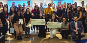 Grande grupo de pessoas reunidas em uma sala de eventos. O grupo é composto por homens e mulheres de diferentes idades e tons de pele, todos posicionados próximos uns dos outros e sorrindo para a câmera, transmitindo um clima de celebração e integração. Na frente, algumas pessoas estão ajoelhadas ou levemente inclinadas, segurando cheques simbólicos de grande tamanho, com valores e textos impressos.