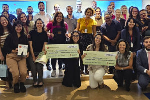 Grande grupo de pessoas reunidas em uma sala de eventos. O grupo é composto por homens e mulheres de diferentes idades e tons de pele, todos posicionados próximos uns dos outros e sorrindo para a câmera, transmitindo um clima de celebração e integração. Na frente, algumas pessoas estão ajoelhadas ou levemente inclinadas, segurando cheques simbólicos de grande tamanho, com valores e textos impressos.