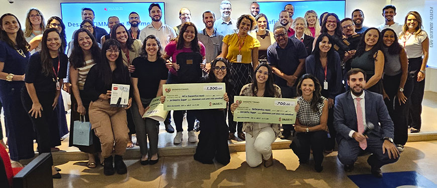 Grande grupo de pessoas reunidas em uma sala de eventos. O grupo é composto por homens e mulheres de diferentes idades e tons de pele, todos posicionados próximos uns dos outros e sorrindo para a câmera, transmitindo um clima de celebração e integração. Na frente, algumas pessoas estão ajoelhadas ou levemente inclinadas, segurando cheques simbólicos de grande tamanho, com valores e textos impressos.