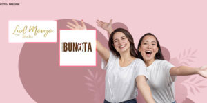 Sob um fundo cor de rosa com flores desenhadas, se vê duas mulheres, ambas de pele clara, cabelos longos castanhos, utilizando calça jeans e blusa branca, com semblante de alegria, na imagem também se vê a logo dos convênios Lud Morgo Studio e Amigas da Bunita.