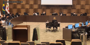 Imagem aberta do auditório do TJMG enquadrando as desembargadoras e os desembargadores membros do Órgão que estão sentados em formação semicircular, lado a lado. As paredes e o carpete do local são em tom marrom e os magistrados usam togas na cor preta.