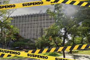 Imagem Acessível: Edifício com fachada em concreto com aberturas formando um padrão de pequenos quadrados, à frente do edifício estão árvores e bancos da Praça Milton Campos. Sobre essa imagem estão faixas zebradas em tons de amarelo e preto com o aviso "SUSPENSO".