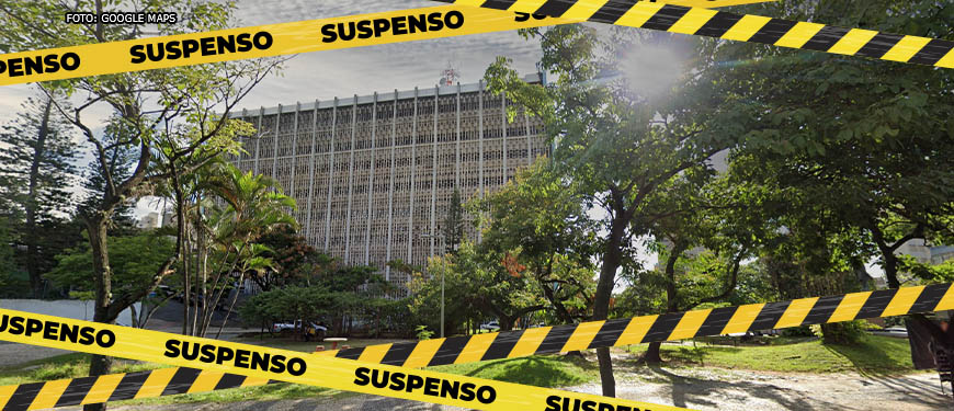 Imagem Acessível: Edifício com fachada em concreto com aberturas formando um padrão de pequenos quadrados, à frente do edifício estão árvores e bancos da Praça Milton Campos. Sobre essa imagem estão faixas zebradas em tons de amarelo e preto com o aviso "SUSPENSO".