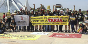 Grupo de manifestantes do SINJUS-MG posa em frente à Catedral de Brasília, durante ato nacional contra a Reforma Administrativa. As pessoas vestem camisetas pretas e seguram faixas com mensagens como “Servidores do Judiciário unidos contra a farsa da Reforma Administrativa” e bandeiras com o logotipo do Sindicato.