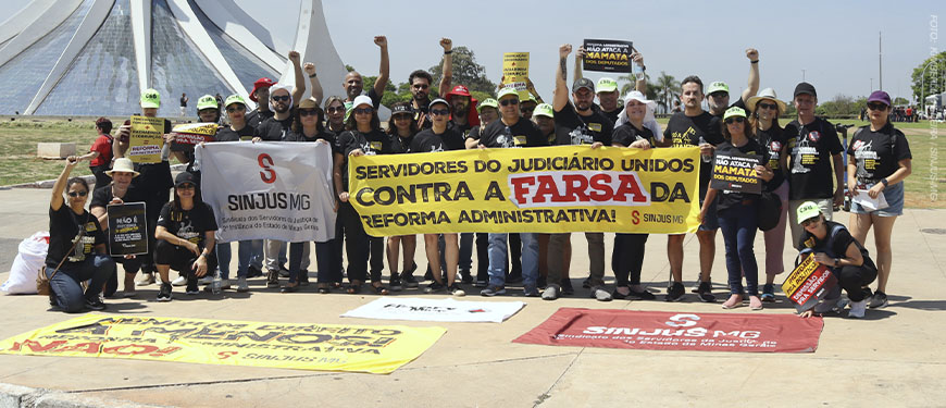 Grupo de manifestantes do SINJUS-MG posa em frente à Catedral de Brasília, durante ato nacional contra a Reforma Administrativa. As pessoas vestem camisetas pretas e seguram faixas com mensagens como “Servidores do Judiciário unidos contra a farsa da Reforma Administrativa” e bandeiras com o logotipo do Sindicato.