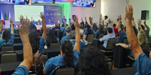 Imagem Acessível: A imagem é um registro fotográfico de um auditório durante o Seminário Intersindical “Inclusão e Dignidade no Serviço Público”, promovido pelo SINJUS-MG. Diversos participantes vestindo camisetas azuis levantam as mãos em sinal de votação ou apoio, enquanto uma mesa de autoridades está disposta à frente, com seis pessoas ao microfone. No telão e nas telas laterais, é exibida a imagem do público presente. O card tem o título “Inclusão e Dignidade” e o texto “SINJUS encaminha ao TJMG pauta dos servidores com deficiência construída durante Seminário”, destacando a luta sindical por acessibilidade e valorização das pessoas com deficiência no Judiciário mineiro