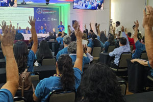 Imagem Acessível: A imagem é um registro fotográfico de um auditório durante o Seminário Intersindical “Inclusão e Dignidade no Serviço Público”, promovido pelo SINJUS-MG. Diversos participantes vestindo camisetas azuis levantam as mãos em sinal de votação ou apoio, enquanto uma mesa de autoridades está disposta à frente, com seis pessoas ao microfone. No telão e nas telas laterais, é exibida a imagem do público presente. O card tem o título “Inclusão e Dignidade” e o texto “SINJUS encaminha ao TJMG pauta dos servidores com deficiência construída durante Seminário”, destacando a luta sindical por acessibilidade e valorização das pessoas com deficiência no Judiciário mineiro