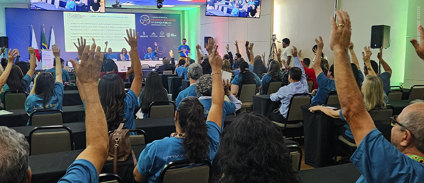 Imagem Acessível: A imagem é um registro fotográfico de um auditório durante o Seminário Intersindical “Inclusão e Dignidade no Serviço Público”, promovido pelo SINJUS-MG. Diversos participantes vestindo camisetas azuis levantam as mãos em sinal de votação ou apoio, enquanto uma mesa de autoridades está disposta à frente, com seis pessoas ao microfone. No telão e nas telas laterais, é exibida a imagem do público presente. O card tem o título “Inclusão e Dignidade” e o texto “SINJUS encaminha ao TJMG pauta dos servidores com deficiência construída durante Seminário”, destacando a luta sindical por acessibilidade e valorização das pessoas com deficiência no Judiciário mineiro