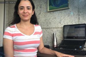 Foto da servidora Érica Borges (mulher de pele clara, cabelos pretos lisos e longos, vestindo uma camisa listrada em branco e rosa) sentada ao lado de um computador da sua estação de trabalho em home office.