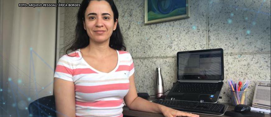 Foto da servidora Érica Borges (mulher de pele clara, cabelos pretos lisos e longos, vestindo uma camisa listrada em branco e rosa) sentada ao lado de um computador da sua estação de trabalho em home office.