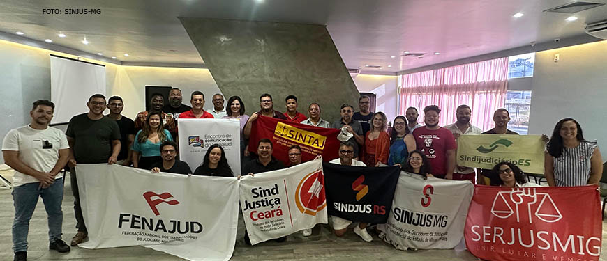 :A imagem mostra um grupo de pessoas reunidas em uma sala, provavelmente para uma reunião ou evento. Eles estão em frente a um palco ou área de apresentação, e alguns indivíduos seguram faixas ou cartazes.