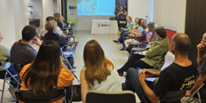 Fotografia de uma palestra realizada em uma sala de reuniões do SINJUS-MG. Cerca de vinte pessoas estão sentadas em cadeiras organizadas em formato semicircular, voltadas para uma tela de projeção. Na tela, há uma apresentação com imagens e texto. Duas mulheres estão em pé na frente da sala, uma delas segurando um microfone e falando com o público. Ao fundo, há uma mesa com o logotipo do SINJUS-MG e banners do NAP e do SINJUS.