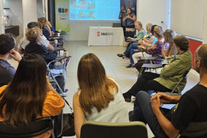 Fotografia de uma palestra realizada em uma sala de reuniões do SINJUS-MG. Cerca de vinte pessoas estão sentadas em cadeiras organizadas em formato semicircular, voltadas para uma tela de projeção. Na tela, há uma apresentação com imagens e texto. Duas mulheres estão em pé na frente da sala, uma delas segurando um microfone e falando com o público. Ao fundo, há uma mesa com o logotipo do SINJUS-MG e banners do NAP e do SINJUS.