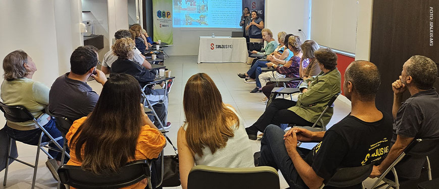 Fotografia de uma palestra realizada em uma sala de reuniões do SINJUS-MG. Cerca de vinte pessoas estão sentadas em cadeiras organizadas em formato semicircular, voltadas para uma tela de projeção. Na tela, há uma apresentação com imagens e texto. Duas mulheres estão em pé na frente da sala, uma delas segurando um microfone e falando com o público. Ao fundo, há uma mesa com o logotipo do SINJUS-MG e banners do NAP e do SINJUS.