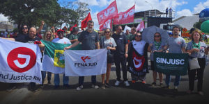 A imagem mostra um grupo de dirigentes e representantes sindicais da FENAJUD e entidades filiadas durante a 2ª Marcha Nacional da Classe Trabalhadora, realizada em Brasília. Eles estão perfilados, segurando bandeiras de sindicatos como o SindJustiça-RN, SINJUSC e da própria FENAJUD.