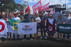 A imagem mostra um grupo de dirigentes e representantes sindicais da FENAJUD e entidades filiadas durante a 2ª Marcha Nacional da Classe Trabalhadora, realizada em Brasília. Eles estão perfilados, segurando bandeiras de sindicatos como o SindJustiça-RN, SINJUSC e da própria FENAJUD.