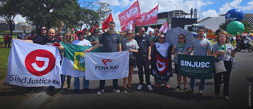 A imagem mostra um grupo de dirigentes e representantes sindicais da FENAJUD e entidades filiadas durante a 2ª Marcha Nacional da Classe Trabalhadora, realizada em Brasília. Eles estão perfilados, segurando bandeiras de sindicatos como o SindJustiça-RN, SINJUSC e da própria FENAJUD.