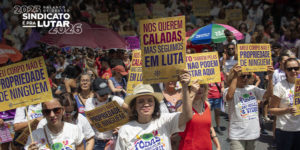 ntegrantes do Núcleo das Mulheres do SINJUS participam da marcha do 8 de Março em meio a uma multidão, segurando cartazes com mensagens de combate à violência e defesa dos direitos das mulheres, durante manifestação em via pública.