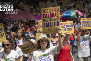 ntegrantes do Núcleo das Mulheres do SINJUS participam da marcha do 8 de Março em meio a uma multidão, segurando cartazes com mensagens de combate à violência e defesa dos direitos das mulheres, durante manifestação em via pública.