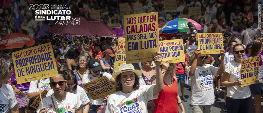 ntegrantes do Núcleo das Mulheres do SINJUS participam da marcha do 8 de Março em meio a uma multidão, segurando cartazes com mensagens de combate à violência e defesa dos direitos das mulheres, durante manifestação em via pública.
