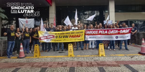 Manifestantes do SINJUS-MG reunidos em frente à sede do TJMG, segurando faixas e bandeiras em defesa da data-base e da valorização dos servidores. Entre os cartazes, destacam-se mensagens como “Unidos pela data-base” e “Se a data-base não chegar, o TJ vai parar”. O grupo ocupa a entrada do prédio em ato organizado pelo sindicato. No canto superior da imagem, aparece a frase “2023: balanço de gestão – Sindicato é pra lutar 2026”.