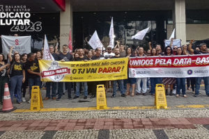Manifestantes do SINJUS-MG reunidos em frente à sede do TJMG, segurando faixas e bandeiras em defesa da data-base e da valorização dos servidores. Entre os cartazes, destacam-se mensagens como “Unidos pela data-base” e “Se a data-base não chegar, o TJ vai parar”. O grupo ocupa a entrada do prédio em ato organizado pelo sindicato. No canto superior da imagem, aparece a frase “2023: balanço de gestão – Sindicato é pra lutar 2026”.