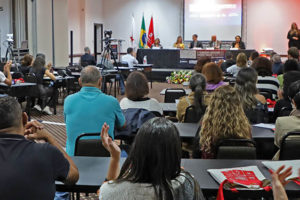 Auditório cheio, a plateia está reagindo com aplausos a uma apresentação. De frente para a plateia, está uma mesa diretiva. Ao fundo e ao centro está um telão, à esquerda dele há um conjunto de bandeiras do Brasil, do estado de Minas Gerais e do SINJUS-MG; do lado direito do telão, está uma intérprete de LIBRAS.