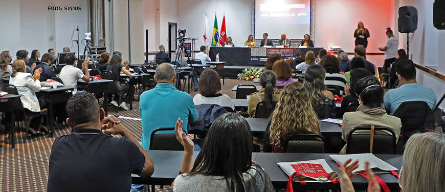 Auditório cheio, a plateia está reagindo com aplausos a uma apresentação. De frente para a plateia, está uma mesa diretiva. Ao fundo e ao centro está um telão, à esquerda dele há um conjunto de bandeiras do Brasil, do estado de Minas Gerais e do SINJUS-MG; do lado direito do telão, está uma intérprete de LIBRAS.