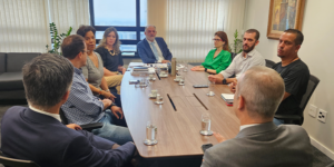 A imagem mostra uma reunião em uma sala com mesa retangular de madeira, com representantes do SINJUS e da 1ª Vice-Presidência do TJMG sentado ao redor da mesa. Todos estão atentos à discussão, com papéis e cadernos sobre a mesa.